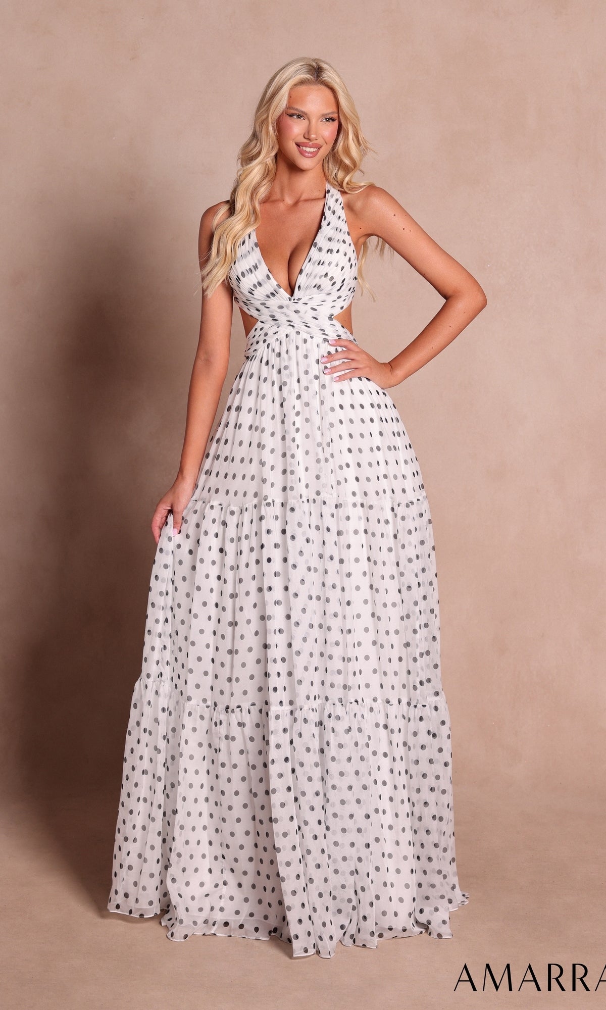 A woman wearing the Amarra 89730 long prom dress in white and black, featuring a halter neckline and chic polka dot print, poses elegantly against a beige background.